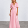 A woman modeling a floor-length metallic pink gown with puff sleeves, a deep V-neckline, and a gathered waist, photographed against a light gray studio backdrop.