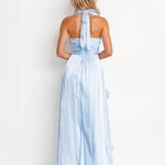 A woman shown from the back wearing a light blue halter-neck maxi dress with a tied back and flowing skirt, photographed against a plain white studio background.