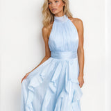 A woman modeling a light blue halter-neck maxi dress with a pleated bodice, a matching waistband, and a layered ruffle skirt, set against a plain white background.
