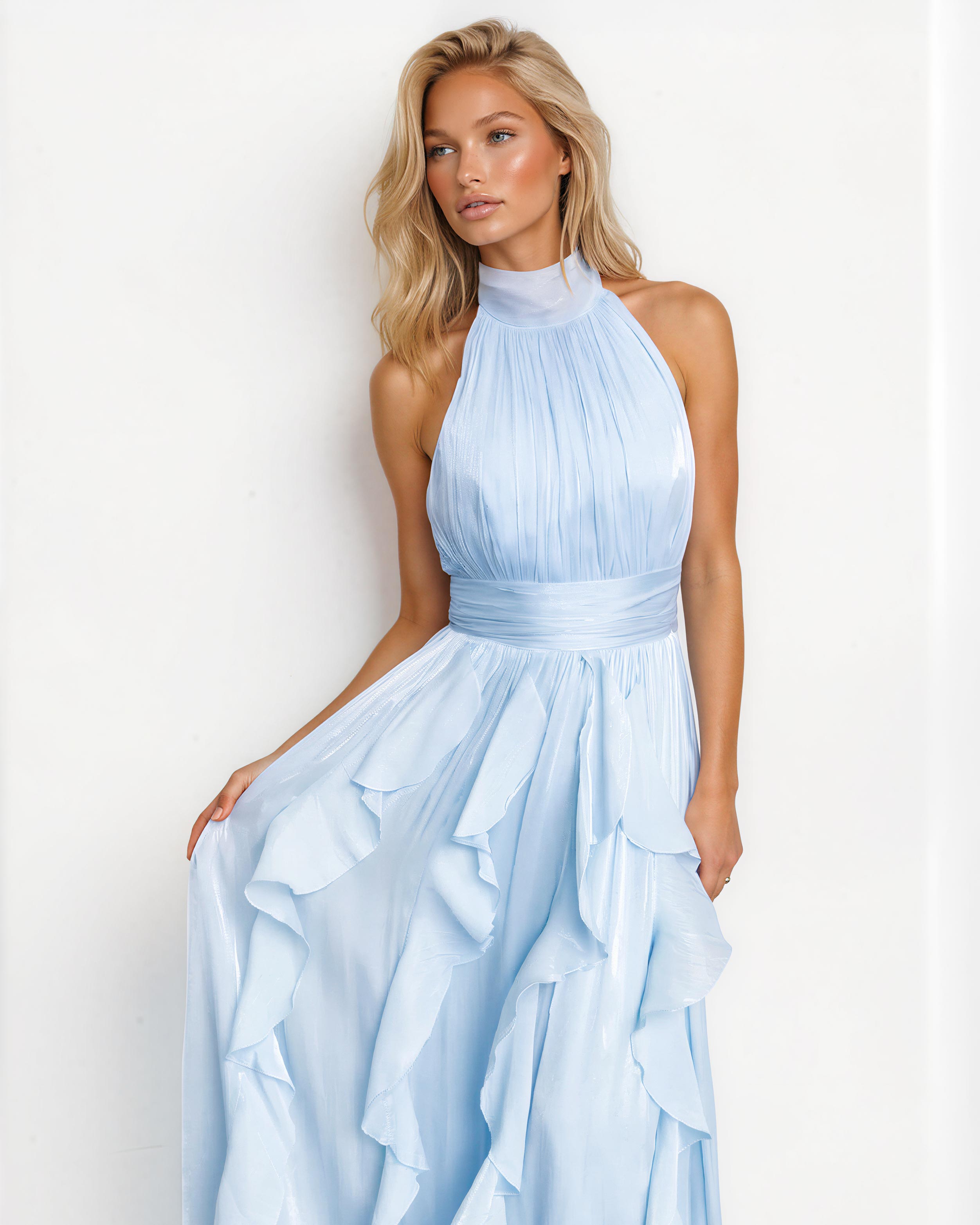 A woman modeling a light blue halter-neck maxi dress with a pleated bodice, a matching waistband, and a layered ruffle skirt, set against a plain white background.