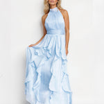 A blonde model wearing a light blue halter-neck maxi dress with a pleated bodice and a layered ruffled skirt, standing against a plain white studio background.