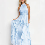 A blonde model wearing a light blue halter-neck maxi dress with a pleated bodice and a layered ruffled skirt, standing against a plain white studio background.