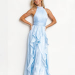 A blonde model wearing a light blue halter-neck maxi dress with a pleated bodice and a layered ruffle skirt, standing against a plain white studio background.