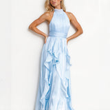 A blonde model wearing a light blue halter-neck maxi dress with a pleated bodice and a layered ruffle skirt, standing against a plain white studio background.