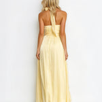 A woman viewed from the back wearing a pale yellow halter-neck maxi dress with a flowing skirt, set against a plain white studio background.