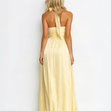 A woman viewed from the back wearing a pale yellow halter-neck maxi dress with a flowing skirt, set against a plain white studio background.