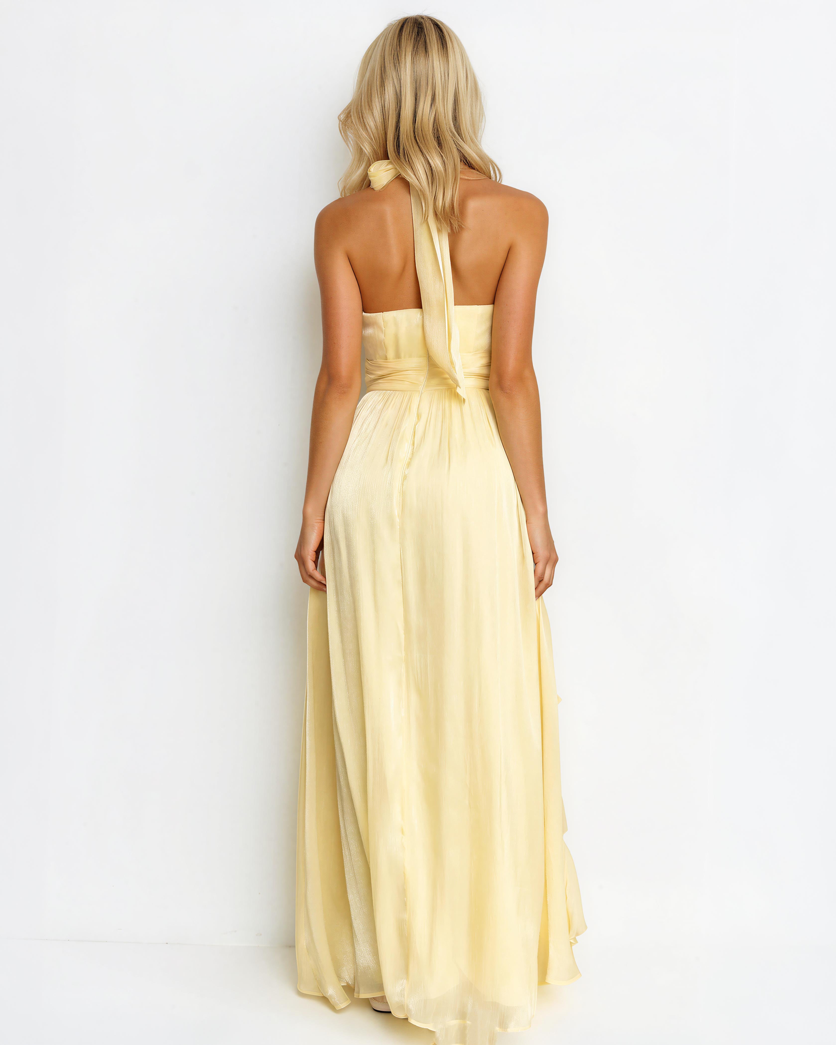 A woman viewed from the back wearing a pale yellow halter-neck maxi dress with a flowing skirt, set against a plain white studio background.