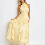 A woman modeling a pale yellow halter-neck maxi dress with a ruffled, tiered skirt, standing against a plain white studio background.