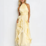 A woman modeling a pale yellow halter-neck maxi dress with a gathered waist and layered ruffle skirt, photographed against a plain white studio background.