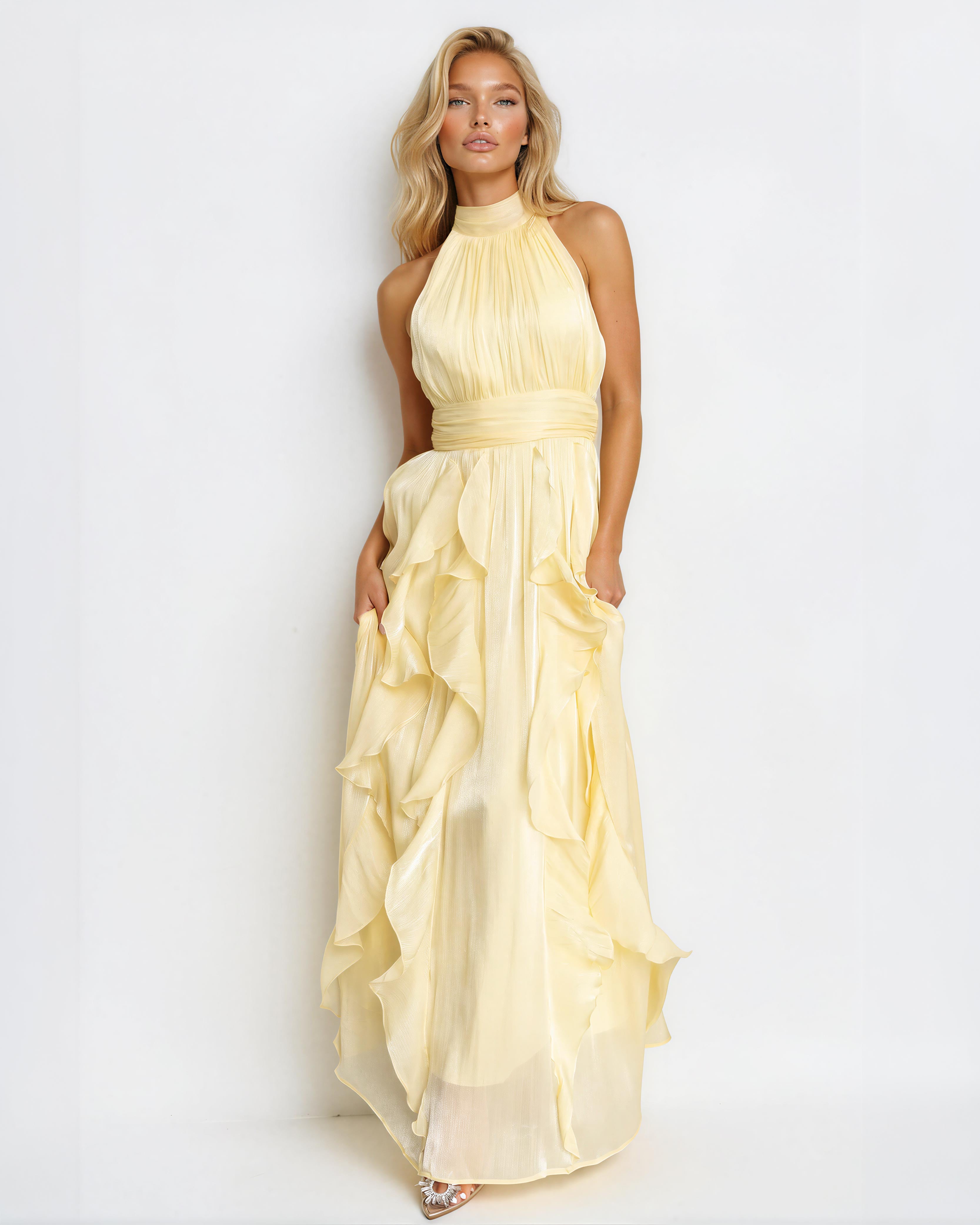 A woman modeling a pale yellow halter-neck maxi dress with a gathered waist and layered ruffle skirt, photographed against a plain white studio background.