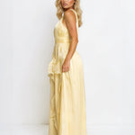 A blonde model wearing a pale yellow sleeveless maxi dress with a ruffled hem, photographed against a plain white studio background.
