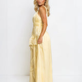 A blonde model wearing a pale yellow sleeveless maxi dress with a ruffled hem, photographed against a plain white studio background.