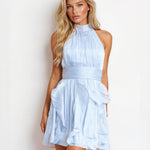 A blonde model wearing a light blue halter-neck mini dress with a pleated bodice and ruffled skirt, posed against a plain white studio background.