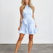 A blonde model wearing a light blue sleeveless halter mini dress with pleated detailing and a ruffled skirt, paired with clear high-heeled sandals, standing against a white studio wall.