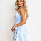 A blonde model wearing a light blue halter-neck mini dress, shown from a side-back angle against a plain white studio background.