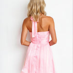 A woman viewed from behind wearing a light pink halter mini dress with a tie-neck detail and a flared skirt, set against a plain white background.