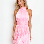 A blonde model wearing a light pink halter-neck mini dress with a ruffled tiered skirt, posed against a plain white studio background.