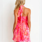 A woman shown from the back wearing a bright pink and coral floral halter mini dress with a tie-neck detail, set against a plain white background.