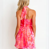 A woman shown from the back wearing a bright pink and coral floral halter mini dress with a tie-neck detail, set against a plain white background.