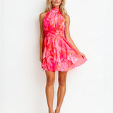 A woman modeling a sleeveless halter-neck mini dress with a vibrant pink and orange tie-dye pattern, standing against a plain white studio background.