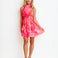 A woman modeling a sleeveless halter-neck mini dress with a vibrant pink and orange tie-dye pattern, standing against a plain white studio background.
