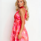 A blonde model wearing a sleeveless halter-neck mini dress with a vibrant pink and orange watercolor floral print, shown from a three-quarter back angle against a plain white background.