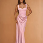 A woman modeling a floor-length light pink satin gown with a sweetheart neckline, structured bodice, and draped skirt against a warm tan studio backdrop.