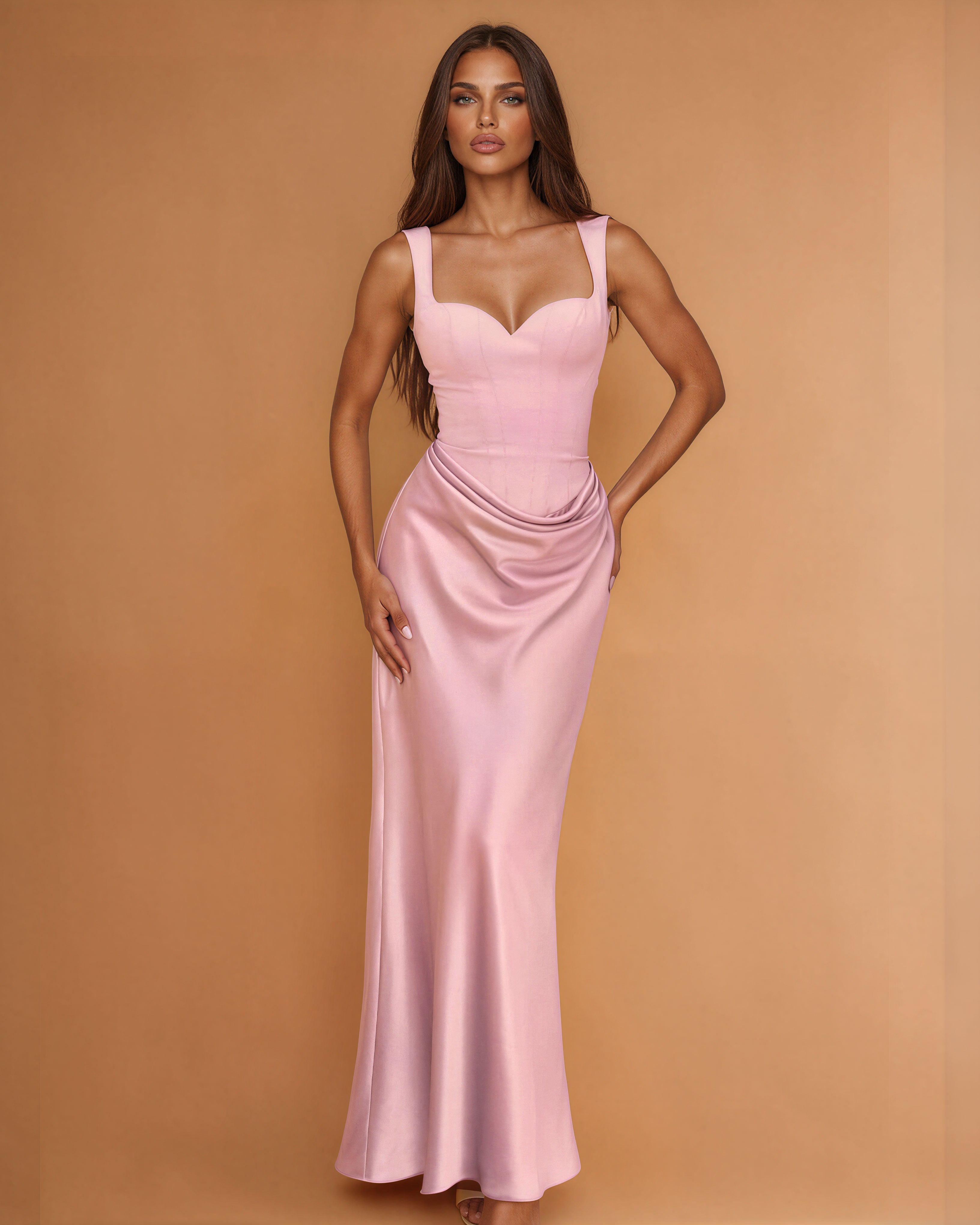 A woman modeling a floor-length light pink satin gown with a sweetheart neckline, structured bodice, and draped skirt against a warm tan studio backdrop.