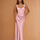 A woman modeling a floor-length light pink satin gown with a sweetheart neckline, structured bodice, and draped skirt against a warm tan studio backdrop.