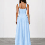 A woman viewed from the back wearing a light blue sleeveless maxi dress with thin shoulder straps and a fitted, flowing skirt, standing against a plain light gray studio background.