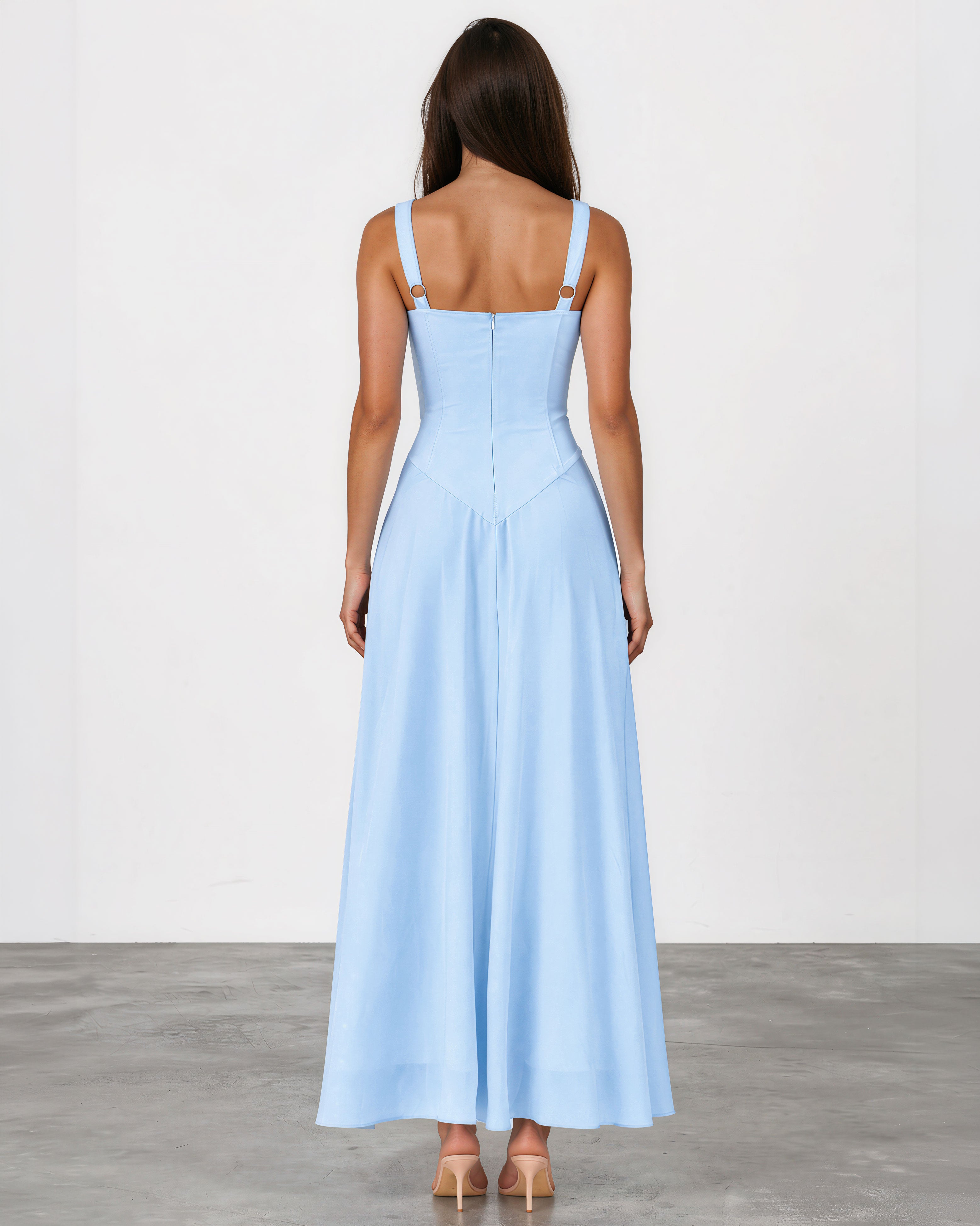 A woman viewed from the back wearing a light blue sleeveless maxi dress with thin shoulder straps and a fitted, flowing skirt, standing against a plain light gray studio background.