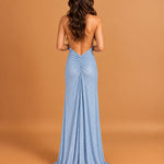 A woman viewed from the back wearing a floor-length light blue evening gown with an open back and gathered waist, standing against a warm tan studio backdrop.