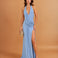 A woman models a light blue halter-neck maxi dress with a deep V-neckline, draped detailing, and a high thigh slit. She is holding a small gold clutch against a solid warm tan studio background.