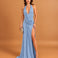 A woman models a light blue halter-neck maxi dress with a draped neckline, a high thigh slit, and a floor-length train against a solid warm tan studio backdrop.