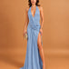 A woman models a light blue halter-neck maxi dress with a draped neckline, a high thigh slit, and a floor-length train against a solid warm tan studio backdrop.