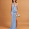 A woman models a light blue glittery halter-neck maxi dress with a draped bodice, thigh-high slit, and a small metallic clutch, set against a solid tan studio background.