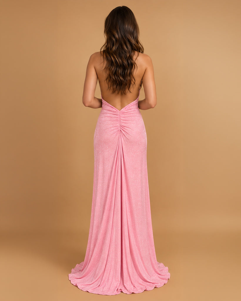 A woman viewed from the back wearing a floor-length pink gown with a draped, gathered back and a slight train, standing against a warm tan studio backdrop.