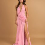A woman modeling a floor-length pink halter gown with a draped wrap-style bodice and a high thigh slit, photographed against a warm tan studio backdrop.