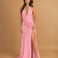A woman modeling a floor-length pink halter gown with a draped wrap-style bodice and a high thigh slit, photographed against a warm tan studio backdrop.