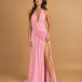 A woman modeling a floor-length pink halter gown with a draped wrap-style bodice and a high thigh slit, photographed against a warm tan studio backdrop.