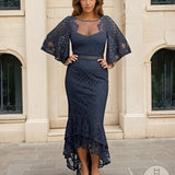 Woman wearing a navy lace dress with a cape in front of a beige building.